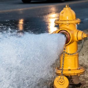 Close-up of yellow fire hydrant gushing water across a street with wet highway and tire from passing car behind